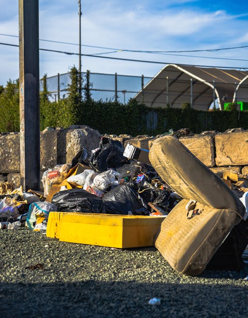 A mound of household rubbish, including black plastic bin bags, cardboard boxes, and miscellaneous waste, is piled on a gravel surface outdoors, near a low stone wall and metal fencing, with a bright