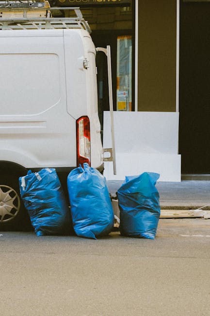 A mound of household rubbish, including black plastic bin bags, cardboard boxes, and miscellaneous waste, is piled on a gravel surface outdoors, near a low stone wall and metal fencing, with a bright