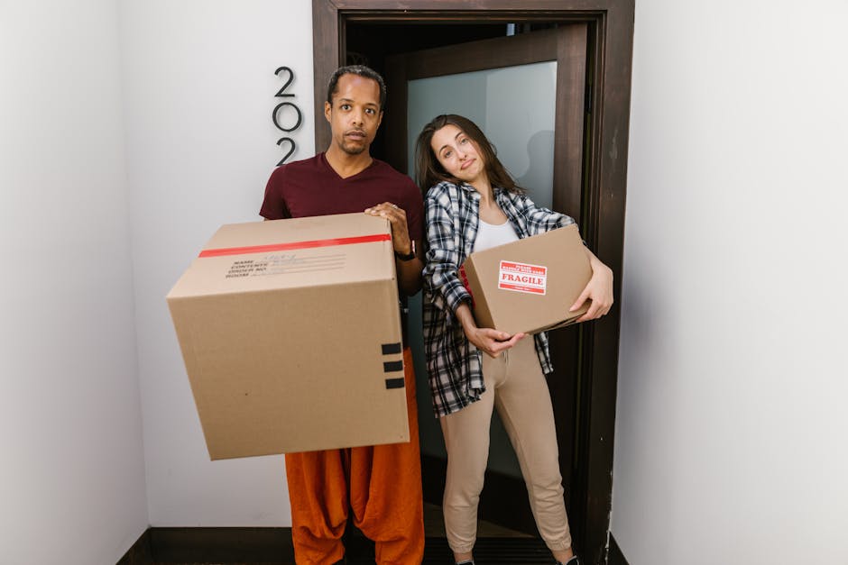 A man and a woman standing inside a residential building near a doorway, both holding cardboard boxes used for home relocation. The man, dressed in a maroon t-shirt and orange trousers, is holding a l