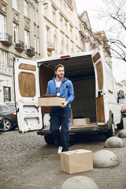 A man and a woman standing inside a residential building near a doorway, both holding cardboard boxes used for home relocation. The man, dressed in a maroon t-shirt and orange trousers, is holding a l