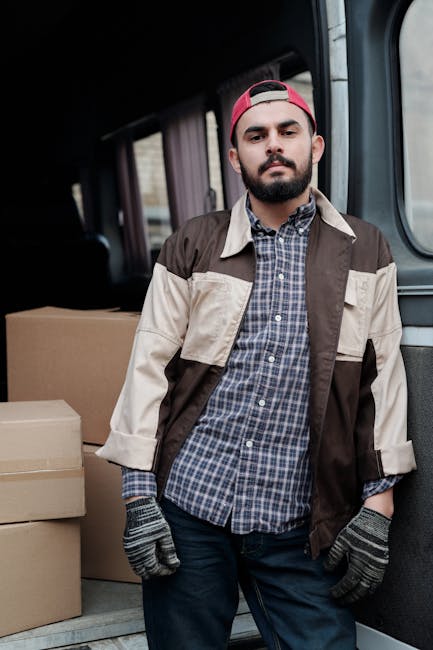 A man wearing a white t-shirt and jeans is standing on a residential street next to an open cargo van with its rear doors swung outward, preparing to load or unload cardboard boxes. The cardboard boxe